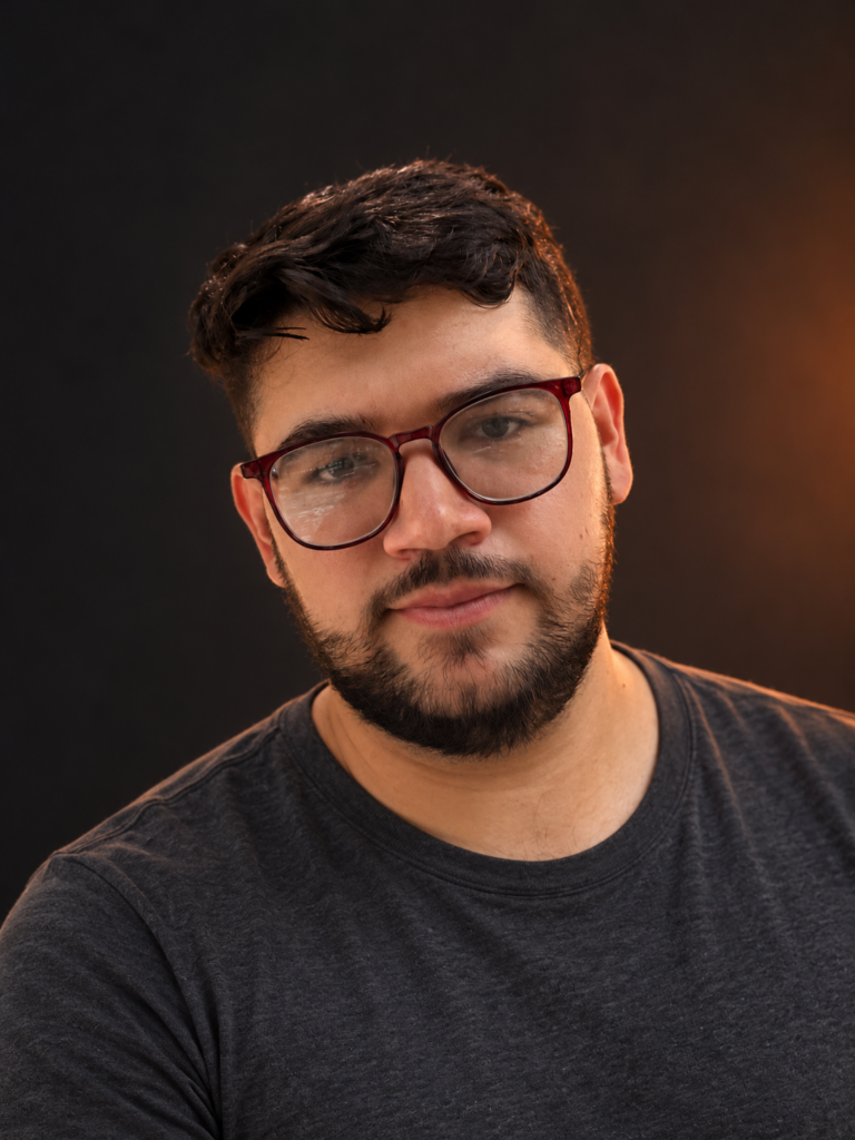Retrato de Fercho Beas con lentes y camiseta casual sobre fondo oscuro.